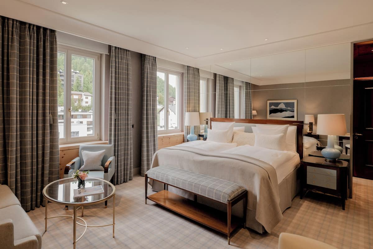 A hotel room with a large bed, two bedside tables with lamps, a small round table with a vase, a bench, and a view of buildings through the windows.