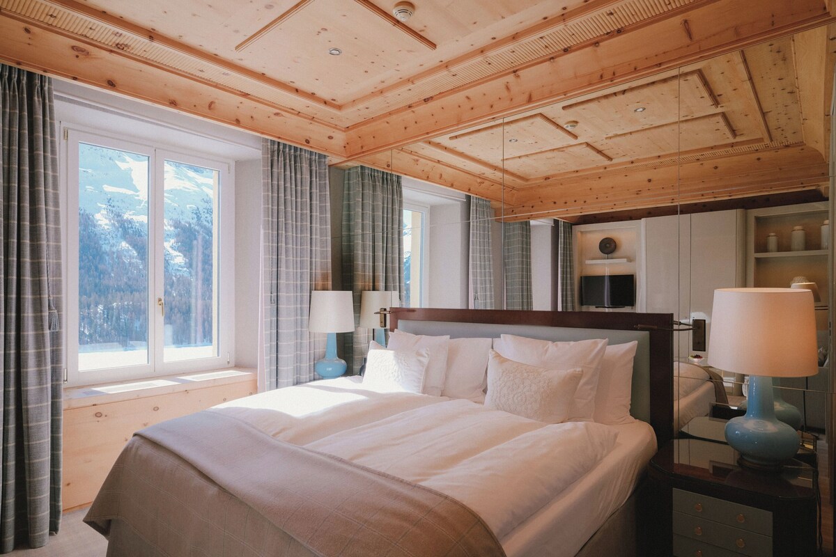 A hotel room with a wooden ceiling, a large bed with white linens, two bedside tables with lamps, and a view of trees through the window.