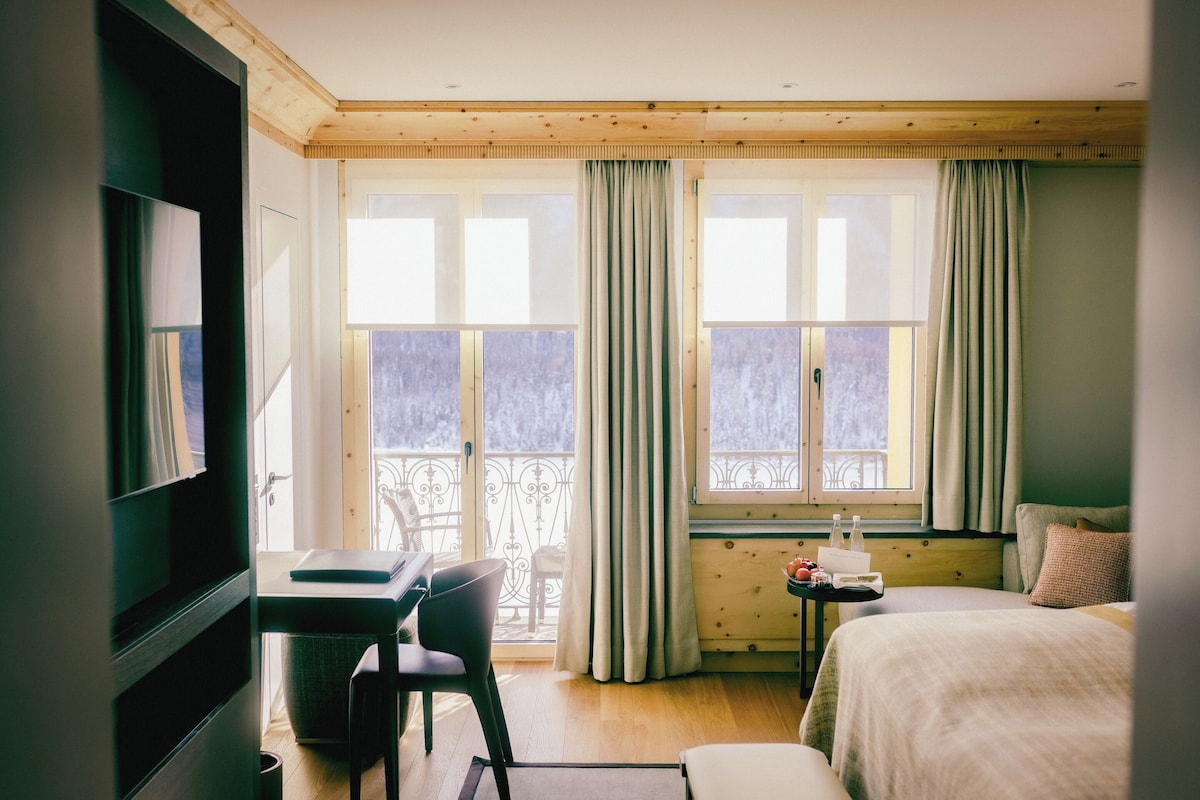A hotel room with a bed, a desk, and a chair. There are two windows with curtains, and a balcony is visible through the windows.