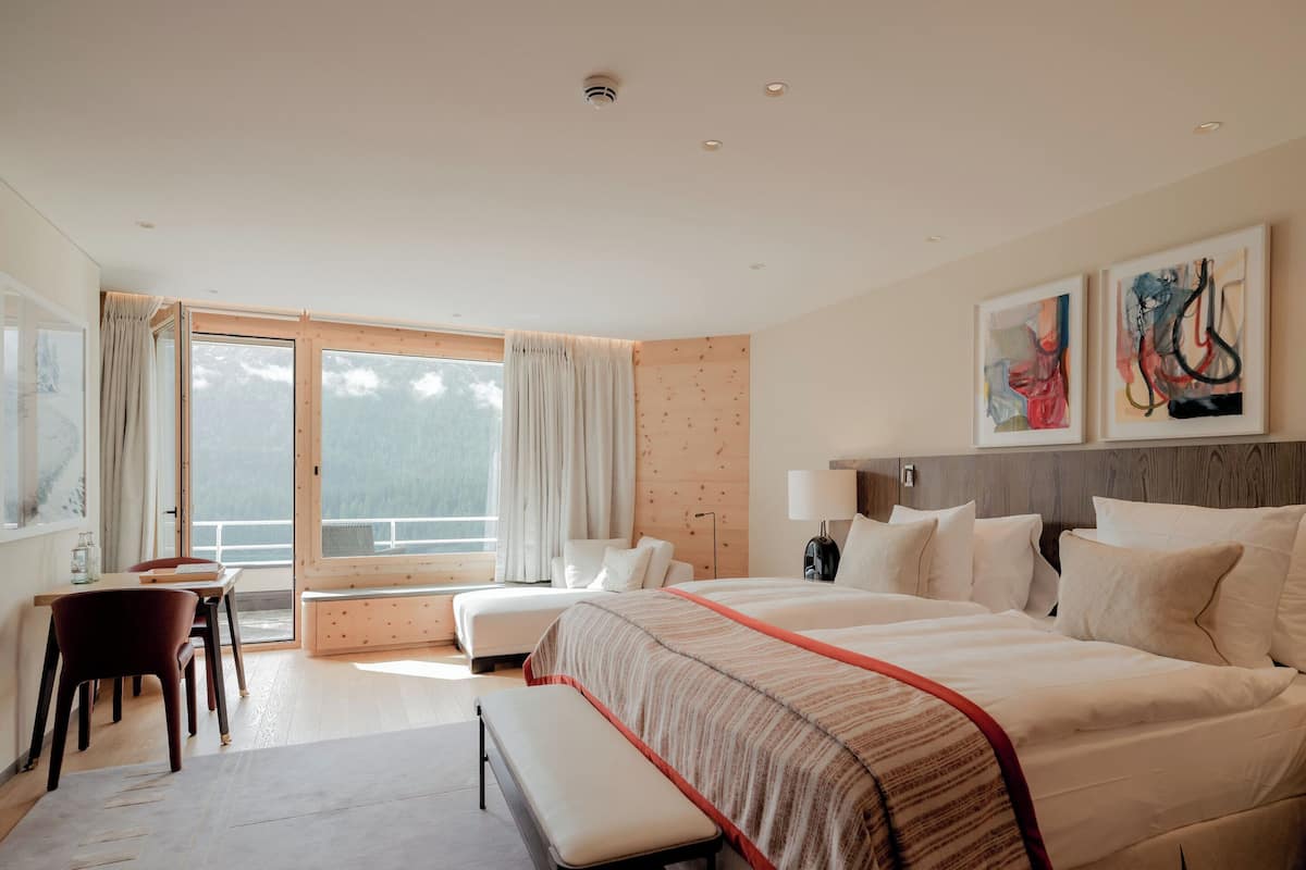 A modern hotel room with a large bed, a desk, and a chair. There is a window with curtains, and artwork on the wall.