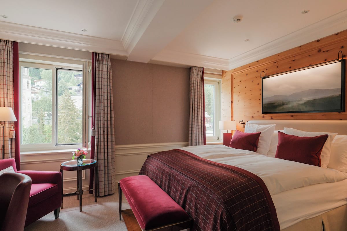 A hotel room with a large bed, a bench, a small table with a vase of flowers, and a painting on the wall.