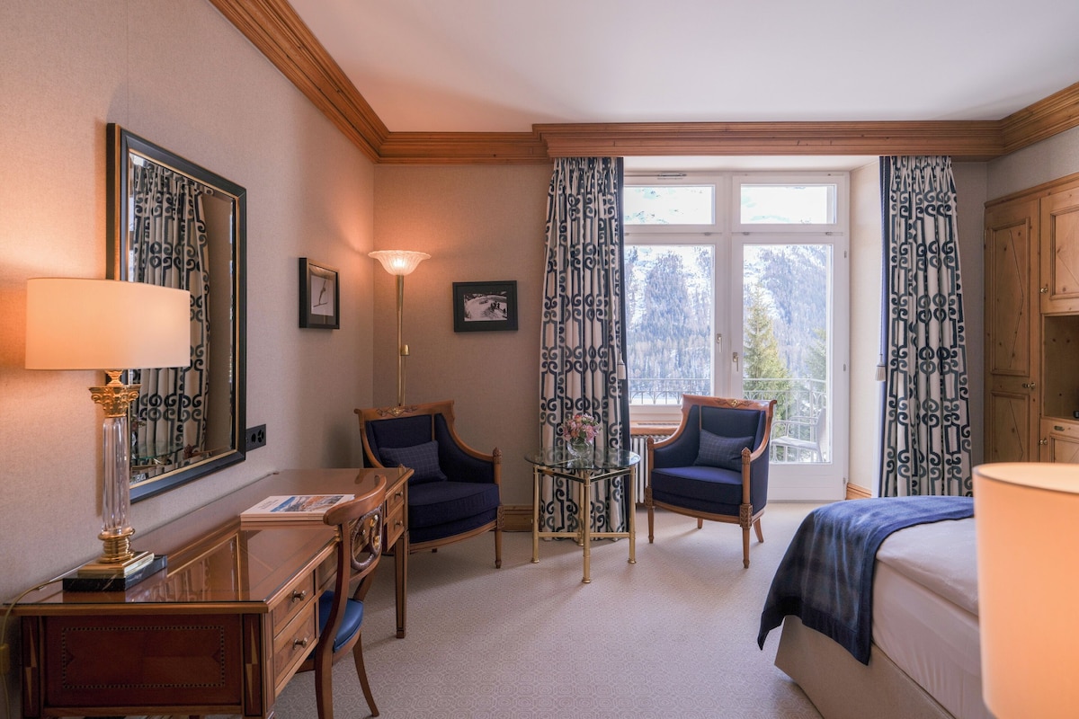 A hotel room with a desk, two chairs, a bed, a large window with curtains, and a mirror.