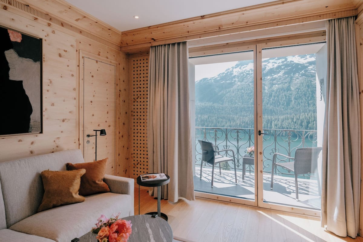 A modern room with a wooden interior, a sofa with cushions, a small table with a vase of flowers, and a balcony with chairs and a view of mountains.