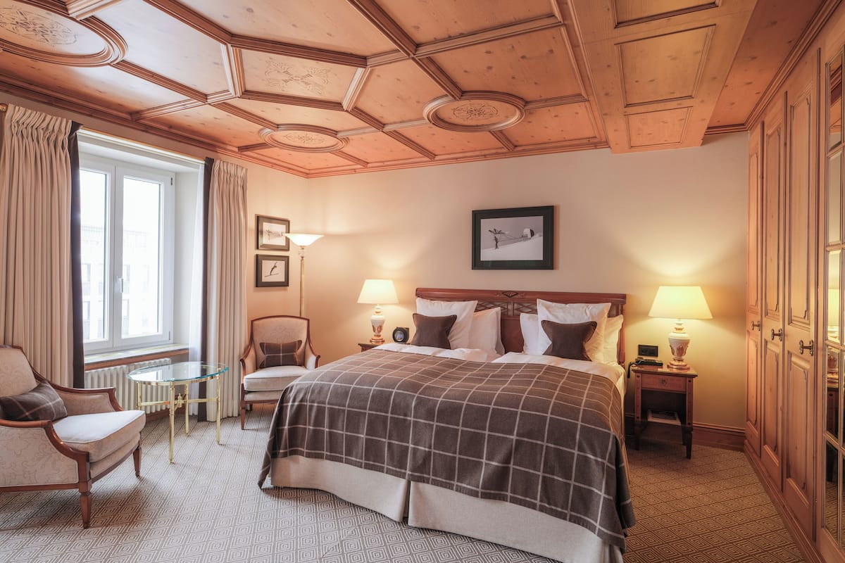 A hotel room with a large bed, two bedside tables with lamps, a chair, a small table, and a window with curtains.