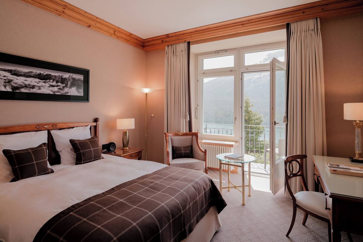 A hotel room with a large bed, a desk, a chair, a small table, and a view of mountains through the window.