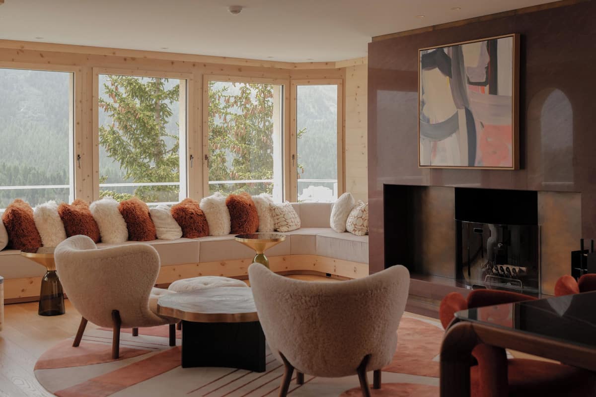 A modern living room with a sofa set, two armchairs, a fireplace, and large windows with a view of trees.