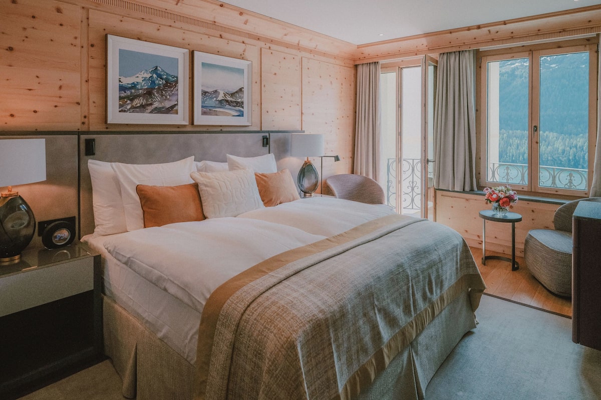 A hotel room with a large bed, a bedside table with a lamp, a small table with flowers, and a view of mountains through the window.