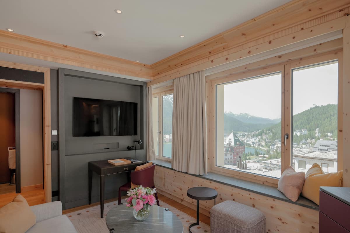 A modern hotel room with a large window offering a city view, a flat-screen TV, a small round table with a vase of flowers, and a cozy seating area.