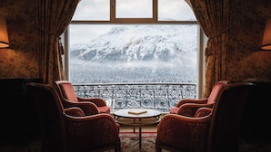 A room with a large window, a balcony with ornate metal railings, and red upholstered chairs. - Kulm Hotel St. Moritz (St. Moritz)