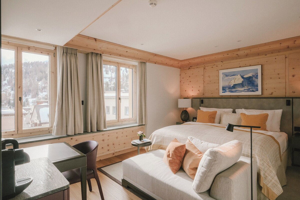 A bedroom with a bed, a desk, and a chair. There is a window with curtains, a painting on the wall, and a view of a snowy landscape.