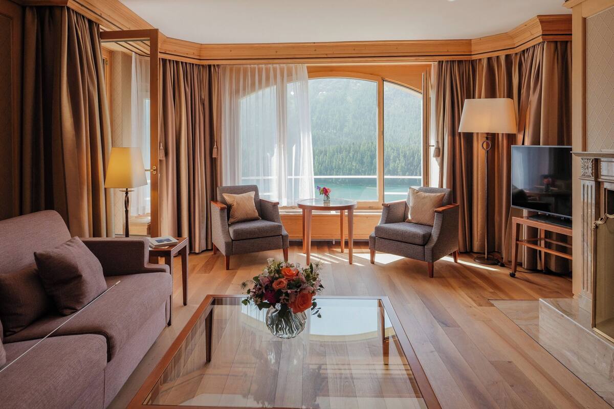 A modern living room with a sofa, two armchairs, a coffee table with flowers, a TV, and a large window with a view of a lake.