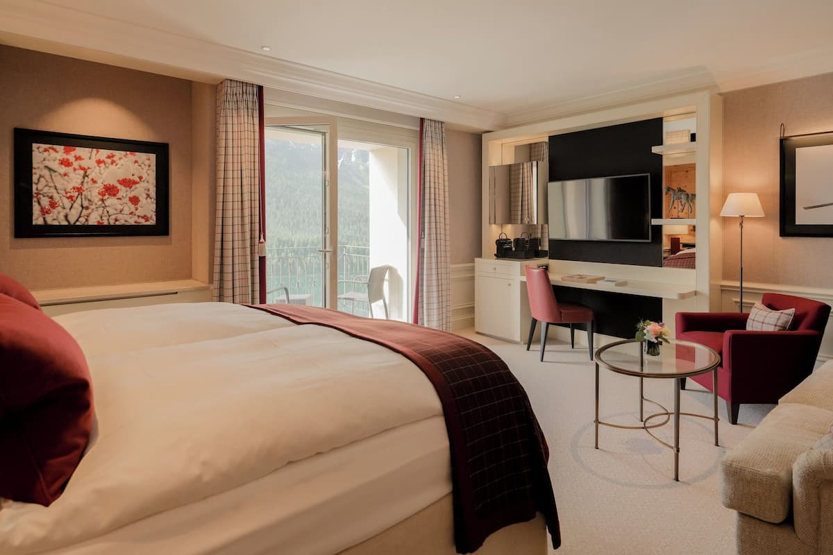 A hotel room with a large bed, a desk, a chair, a TV, and a balcony with a view of the mountains.