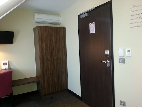 A room with a wooden wardrobe, a desk with a chair, a mounted TV, and a door with a lock and a control panel.