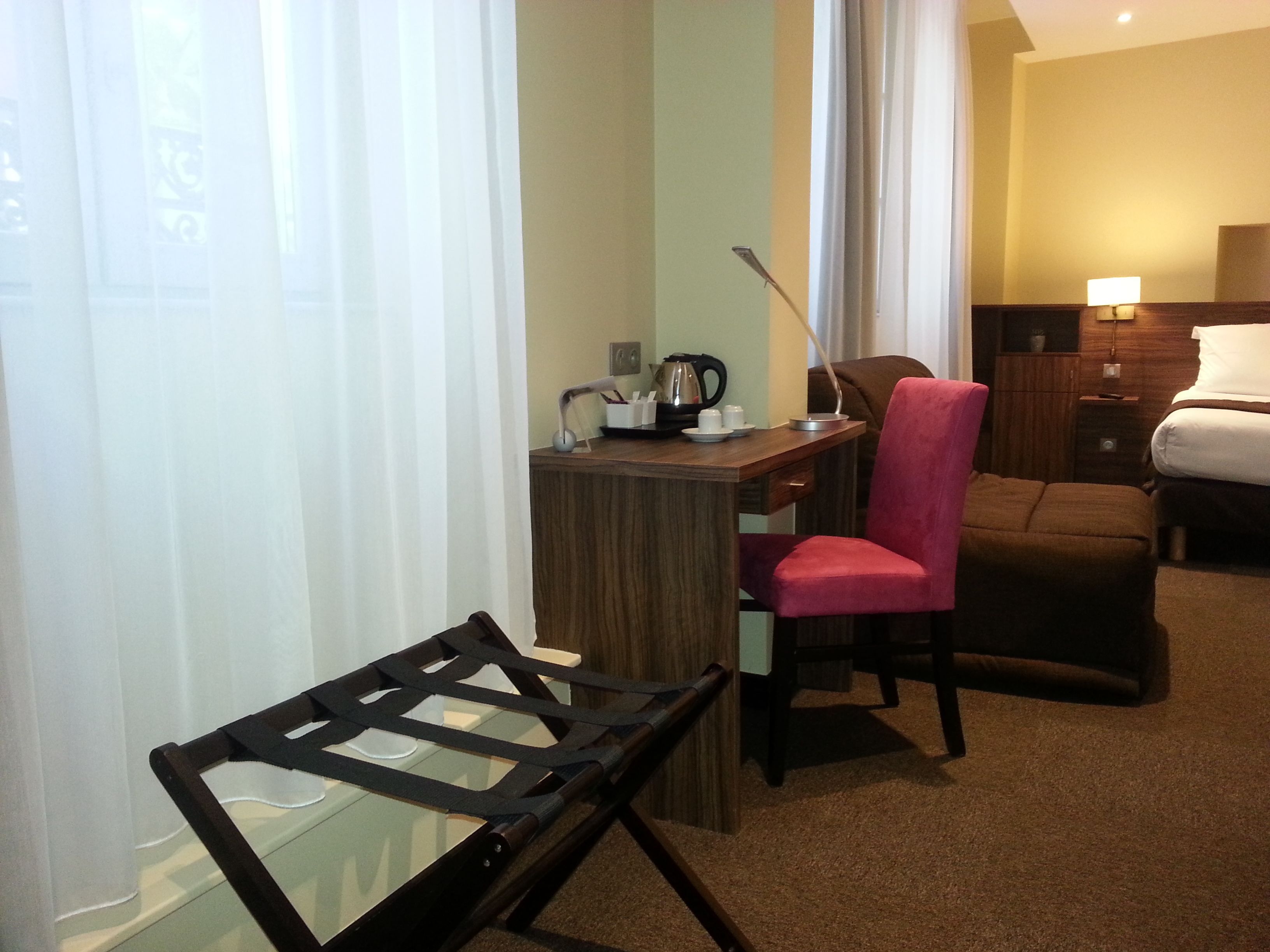 A hotel room with a desk, chair, bed, and a small table with a lamp.