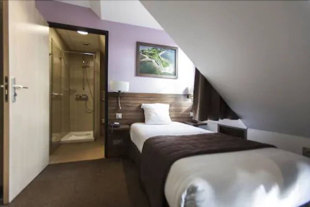 A hotel room with a bed, a bedside table, a nightstand, a lamp, a painting on the wall, and a shower area.