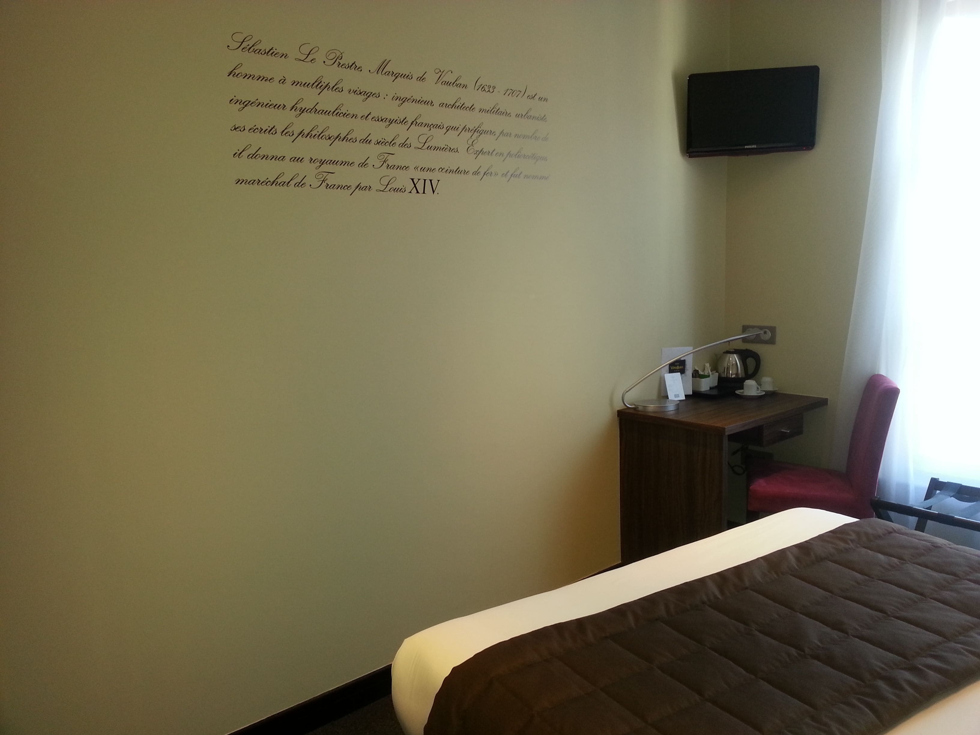 A hotel room with a bed, a desk with a chair, a flat-screen TV mounted on the wall, and a window with curtains.