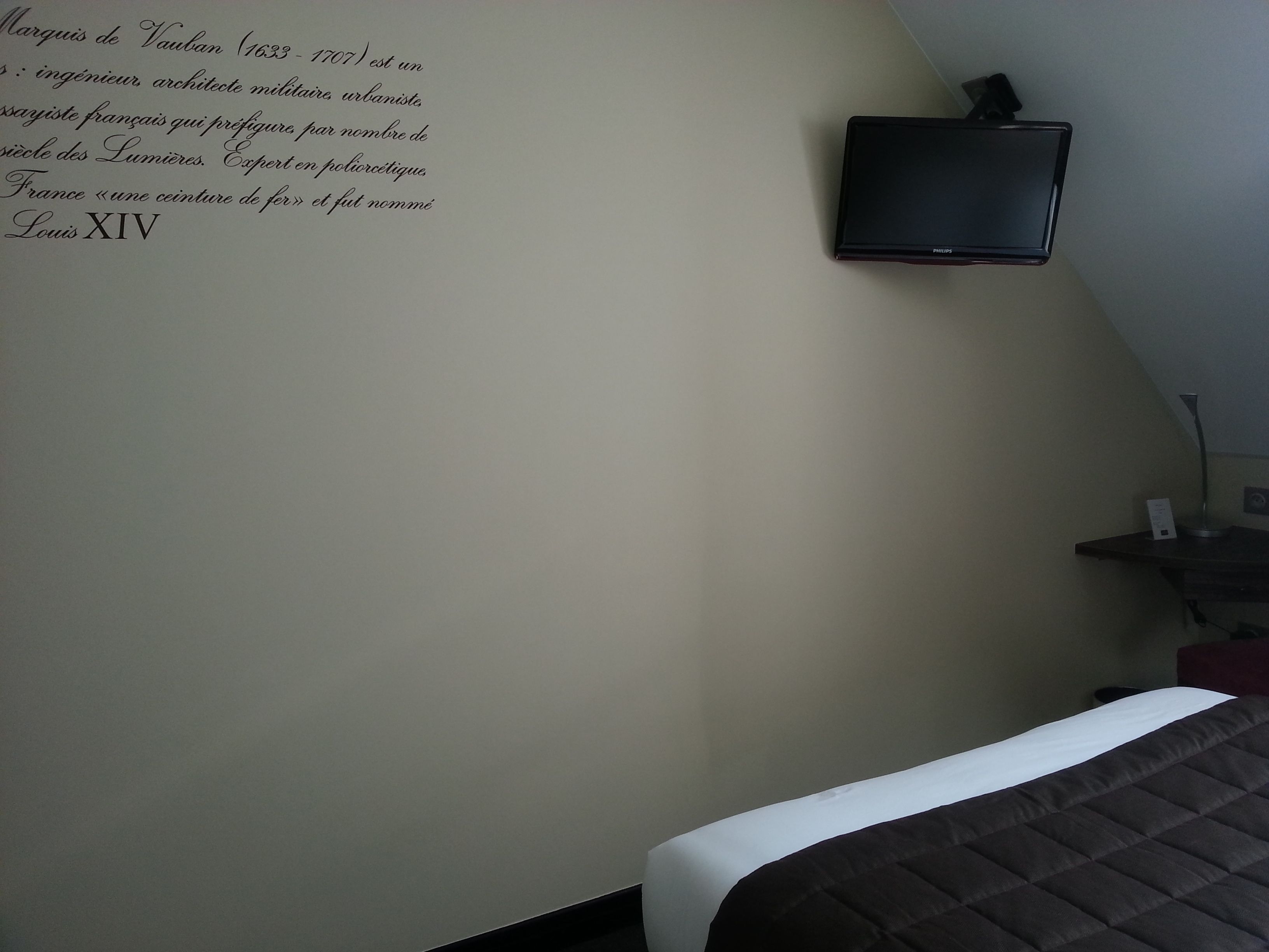 A hotel room with a bed, a flat-screen TV mounted on the wall, and a small table with a lamp.