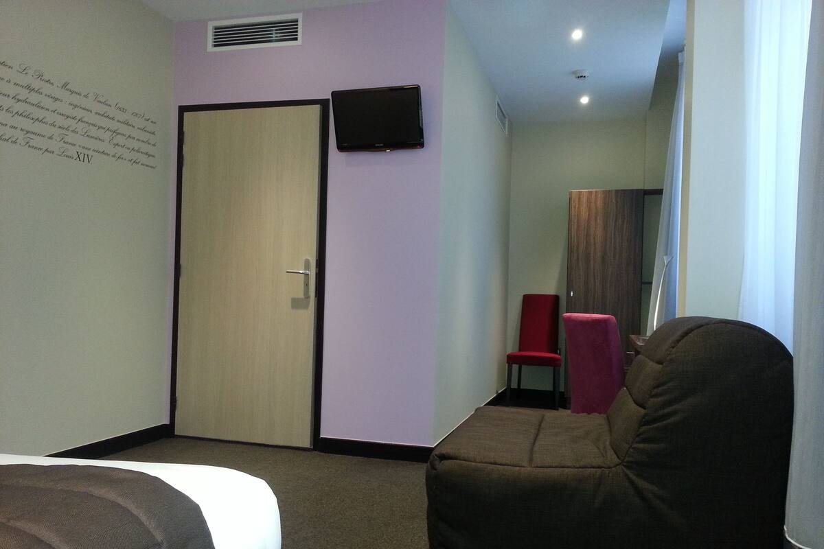 A hotel room with a bed, a red chair, a brown sofa, a television on the wall, and a door.