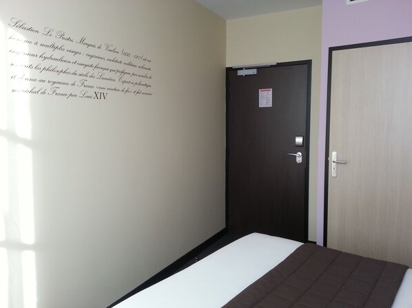 Un pasillo estrecho con una cama al final, una puerta con un cartel y una pared con texto.