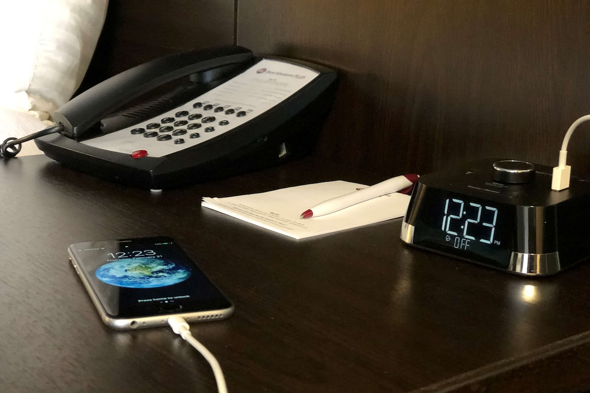 Une chambre d’hôtel équipée d’un bureau comprenant un téléphone à fil, une horloge numérique, un smartphone et un bloc-notes.