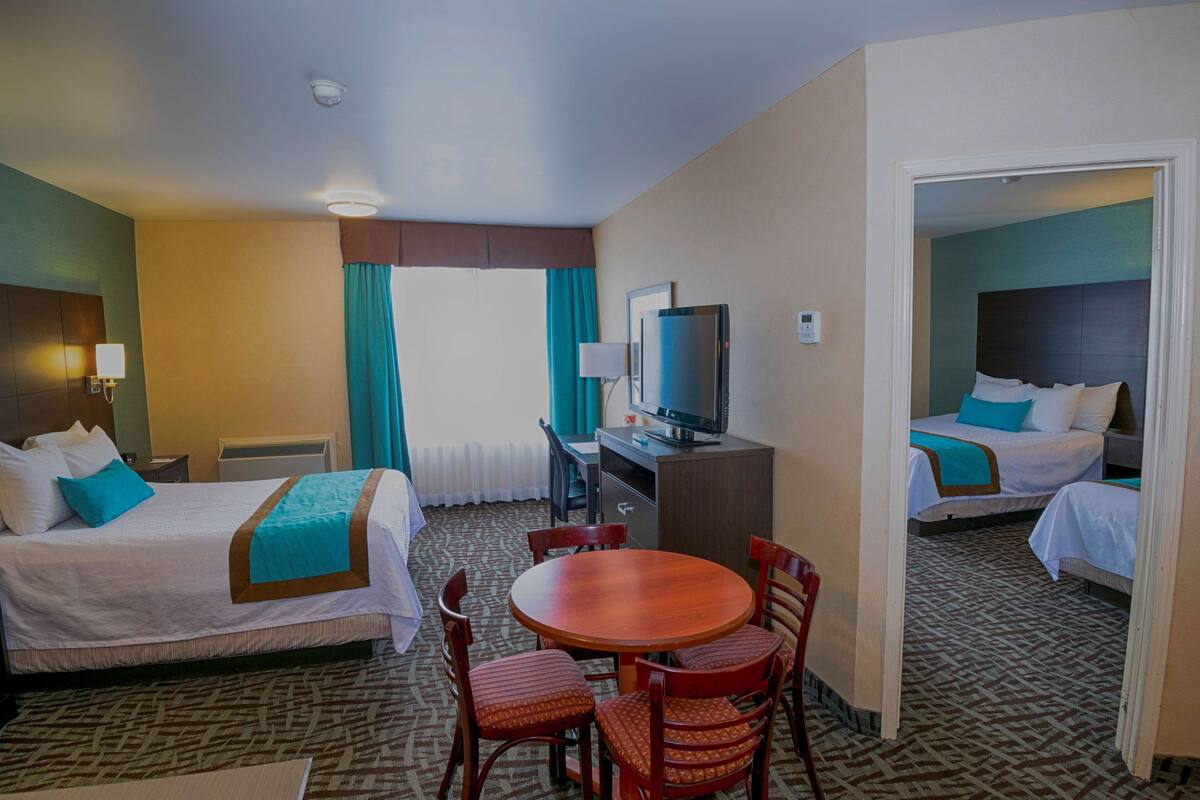 A hotel room with two beds, a desk with a television, a dining table with chairs, and a patterned carpet.
