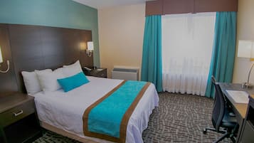 Habitación de hotel con cama, escritorio, silla, mesita de noche, lámpara y ventana con cortinas.