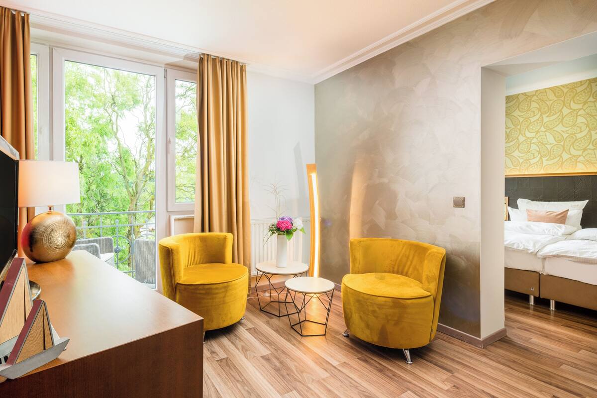 Une chambre d’hôtel avec un plancher en bois, deux fauteuils jaunes, une petite table ronde, un lit avec une literie blanche et une grande fenêtre avec des rideaux.