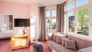 A hotel room with a bed, a sofa, a TV, a wooden nightstand, a grey ottoman, and a window with a view of the outdoors. - Hotel Residenz Waldkrone (Kuehlungsborn)