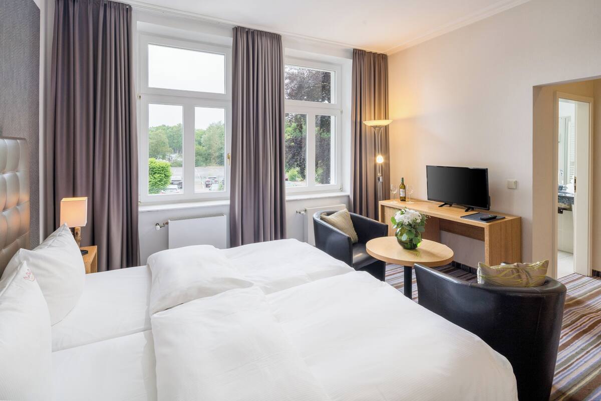 Une chambre d’hôtel avec un grand lit, un bureau avec une télévision, une chaise et une petite table sur laquelle est posé un vase de fleurs.