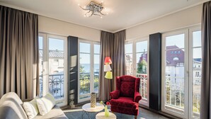 A modern hotel room with a sofa, a red armchair, two small round tables, and large windows offering a view of the sea and buildings. - Hotel Residenz Waldkrone (Kuehlungsborn)