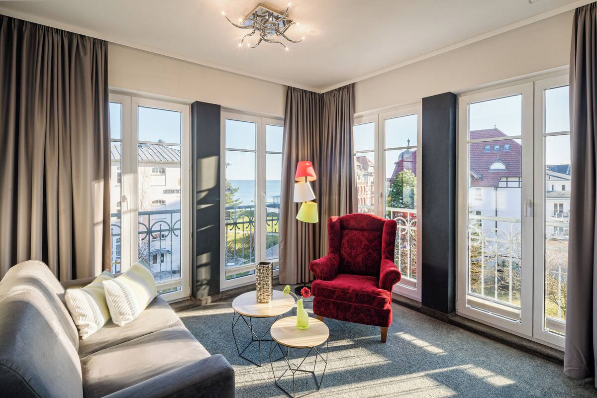 Une chambre d’hôtel moderne, dotée d’un canapé, d’un fauteuil rouge, de deux petites tables rondes et de grandes fenêtres offrant une vue sur la mer et les bâtiments.