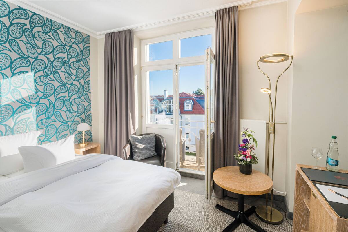 Une chambre d’hôtel comprenant un lit, un bureau, une chaise, une lampe sur pied, une fenêtre avec des rideaux et une vue sur les bâtiments environnants.