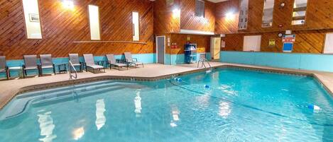 An indoor swimming pool with wooden walls and a tiled floor. There are lounge chairs and a changing room visible.
