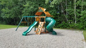 A playground with a wooden play structure, including a slide and climbing area, set on a gravel surface with trees in the background. - Eastern Inn & Suites (North Conway)