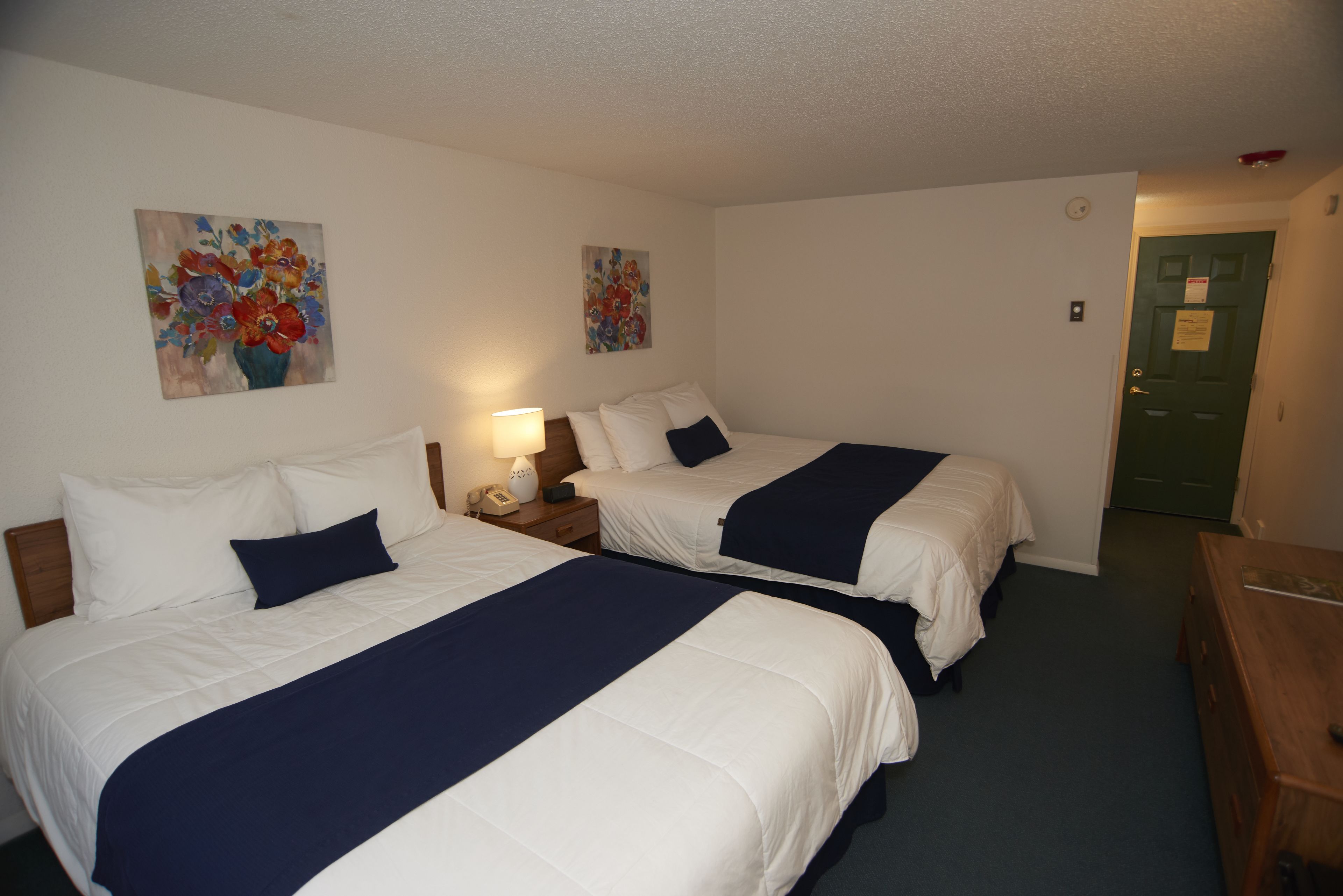 A hotel room with two beds, each with white bedding and navy blue throw pillows. There are two paintings on the walls, a wooden nightstand with a lamp, and a green door.
