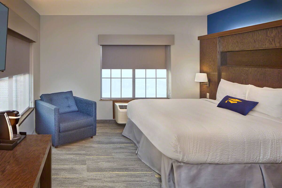 A hotel room with a bed, a blue armchair, a window with blinds, a wall-mounted lamp, and a flat-screen TV.