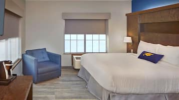 Habitación de hotel con una cama, un sillón azul, una ventana con persianas, una lámpara de pared y un televisor de pantalla plana.