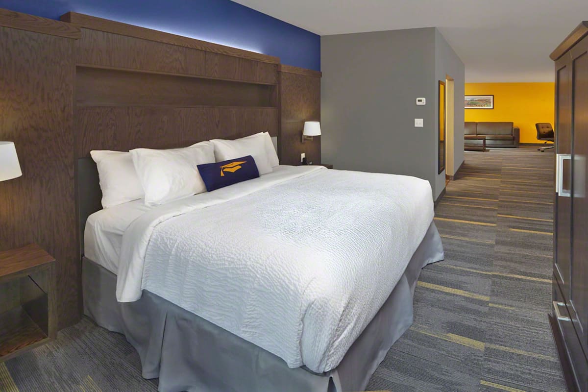 A hotel room with a large bed, a wooden headboard, a nightstand, a lamp, and a view of a hallway with a couch and a chair.