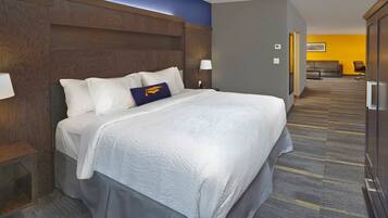 A hotel room with a large bed, a wooden headboard, a nightstand, a lamp, and a view of a hallway with a couch and a chair.