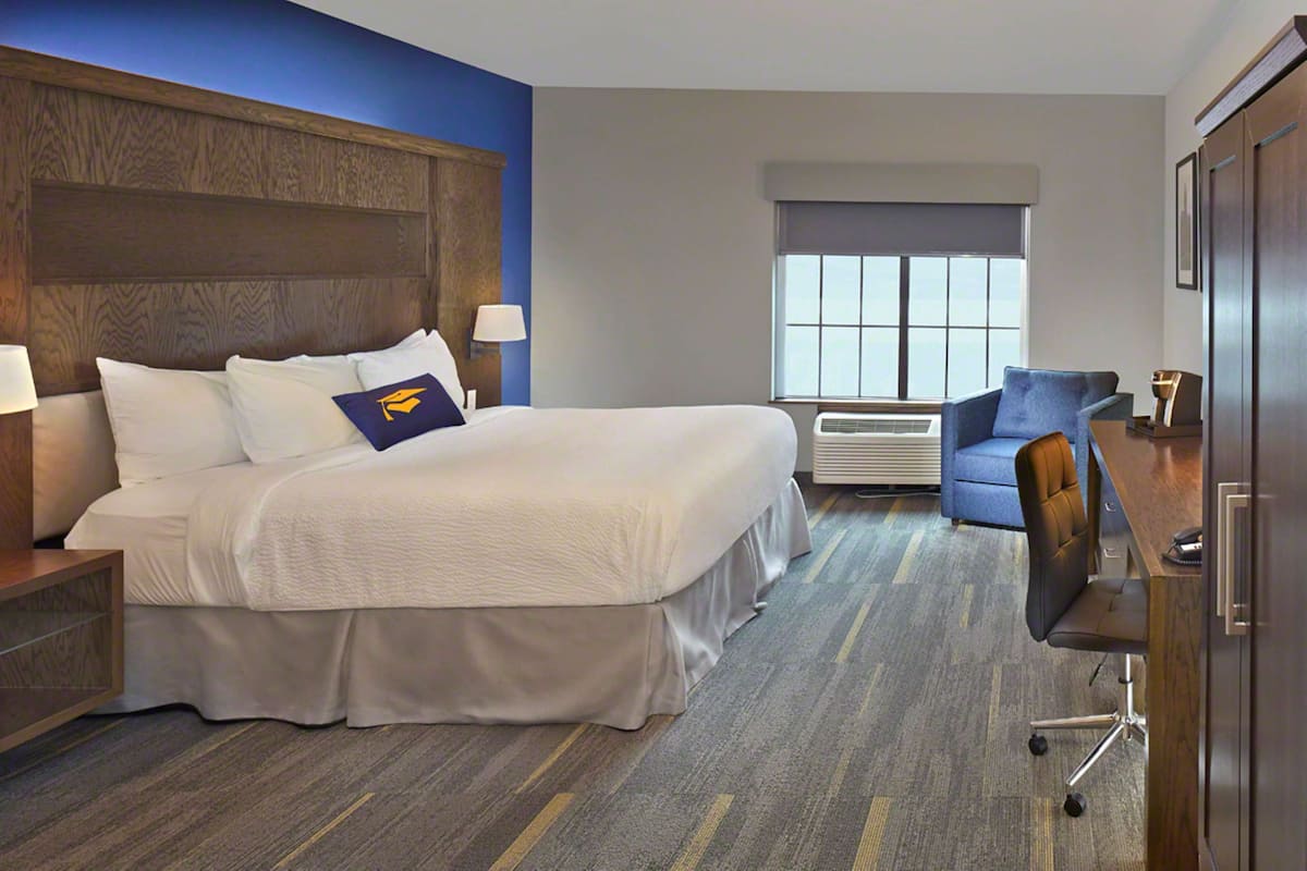 A hotel room with a large bed, a desk, a chair, a window with blinds, and a wooden headboard.