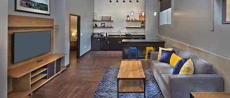 Un salón moderno con televisor de pantalla plana, mesa de centro de madera y sofá con cojines estampados. Se ve un área de cocina al fondo, con un taburete de bar y una lámpara colgante.