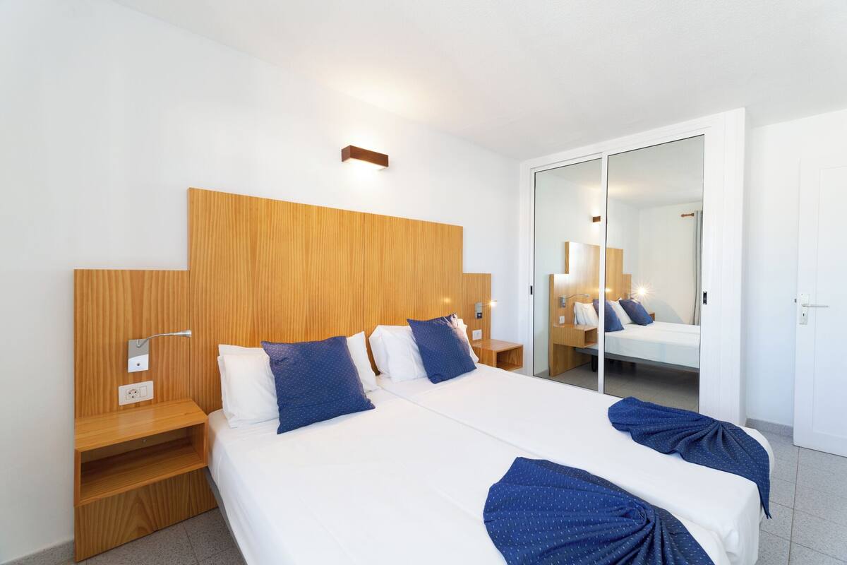 A modern hotel room with a large bed, wooden headboard, bedside tables, and a mirror.