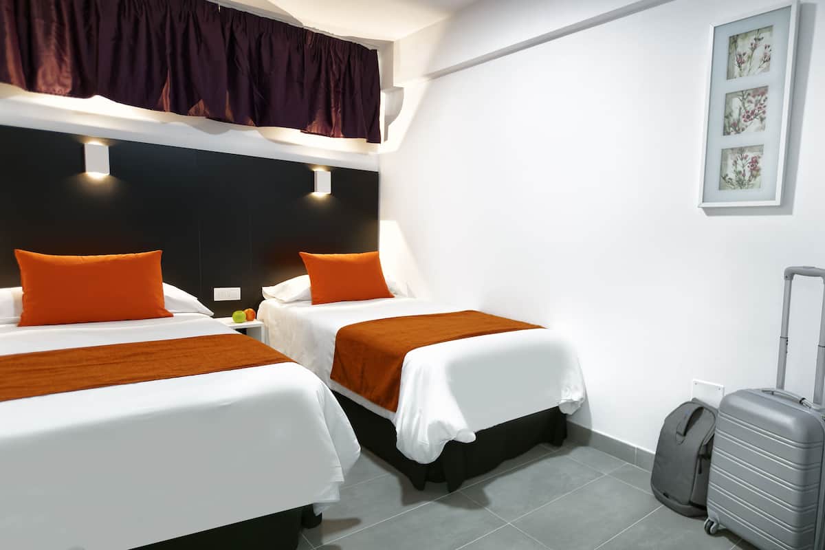 Ein Hotelzimmer mit zwei Betten, jeweils mit weißer Bettwäsche und orangefarbenen Kissen, einem dunklen Kopfteil und einem großen Fenster mit dunklem Vorhang.