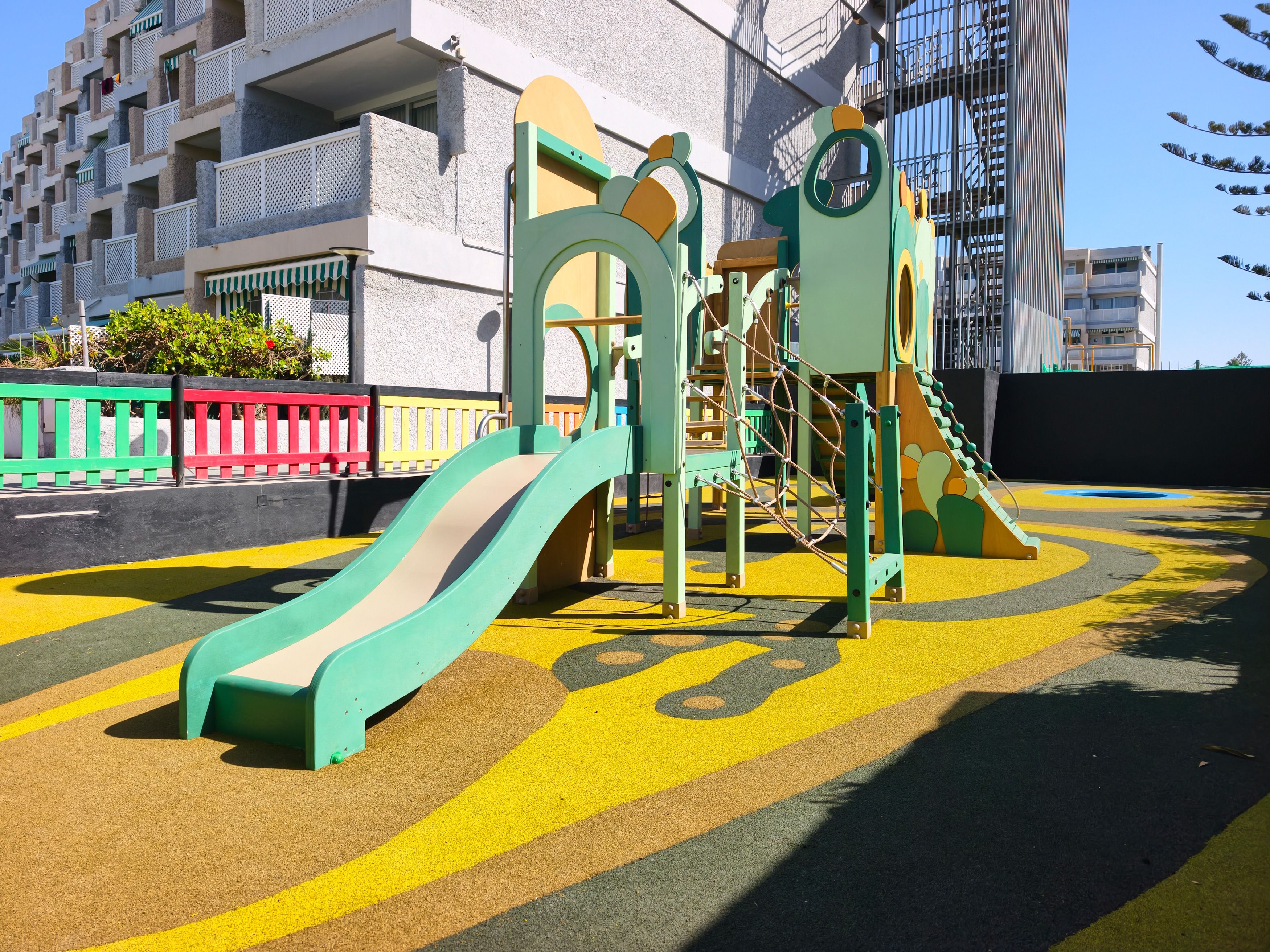 A playground with a slide and climbing structures, surrounded by a colorful fence and a building in the background.