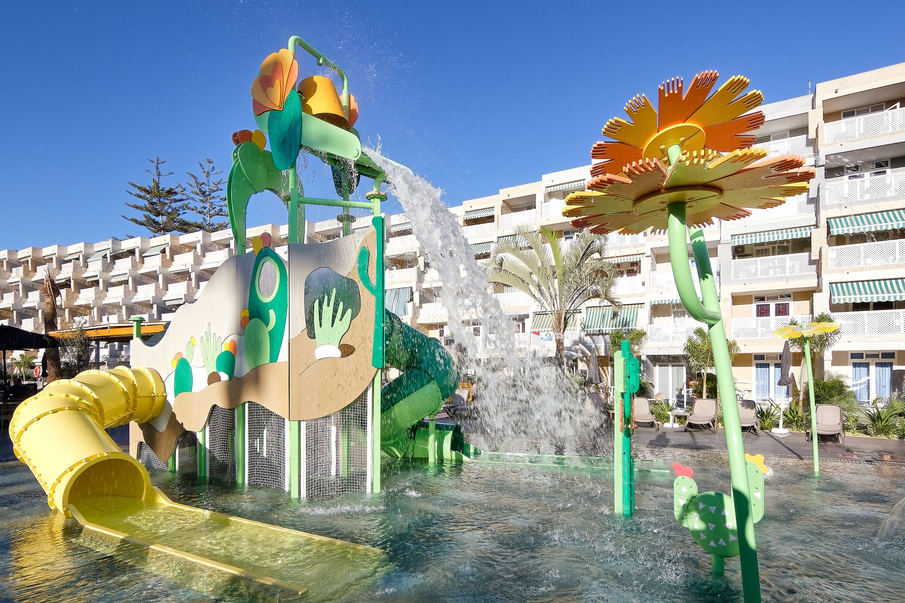 A water play area with slides and fountains, surrounded by a building with balconies and outdoor seating.