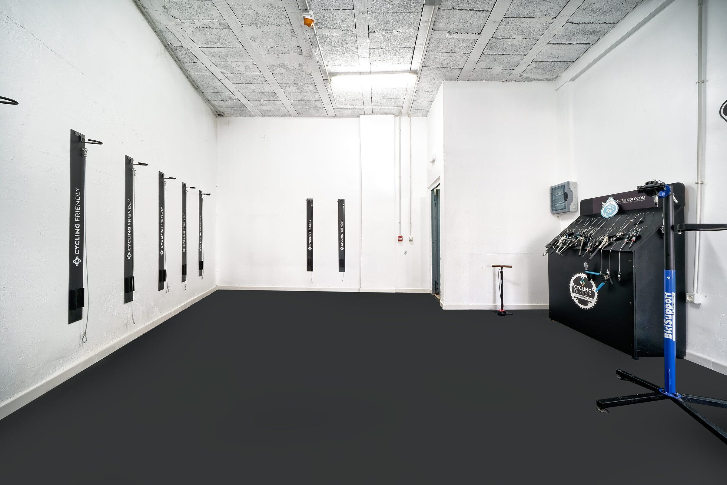 Habitación con paredes blancas, piso negro y techo con vigas a la vista. Se observan barras verticales negras en la pared, una máquina de ejercicio negra y una pequeña mesa con una silla.