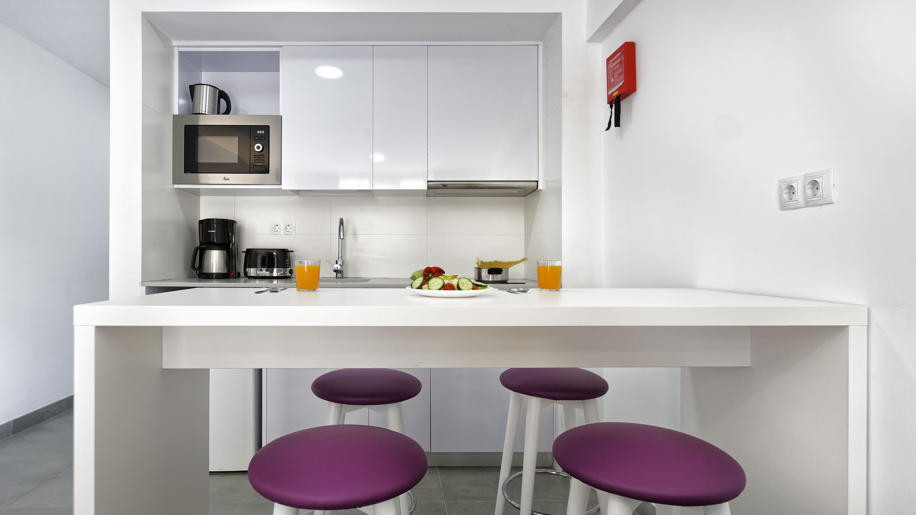 A modern kitchen with white cabinets, a microwave, a coffee maker, and a sink. There are purple stools at the counter.