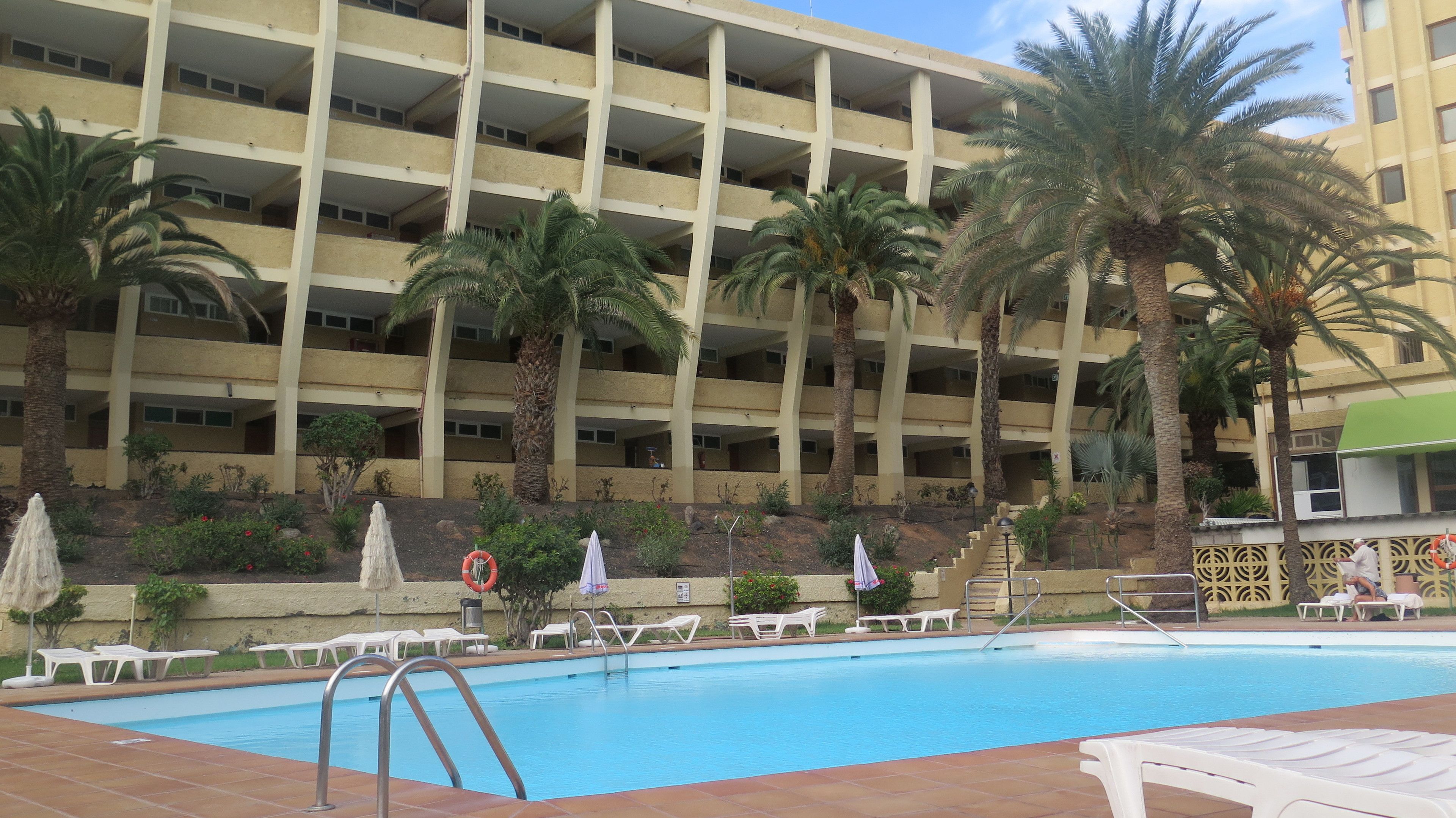 A swimming pool with lounge chairs and umbrellas, surrounded by palm trees and a multi-story building with balconies.