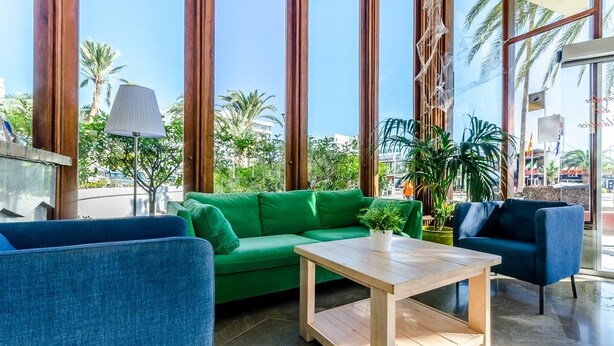 A modern lobby with a green sofa, blue armchairs, and a wooden coffee table. Large windows offer views of palm trees and buildings outside.