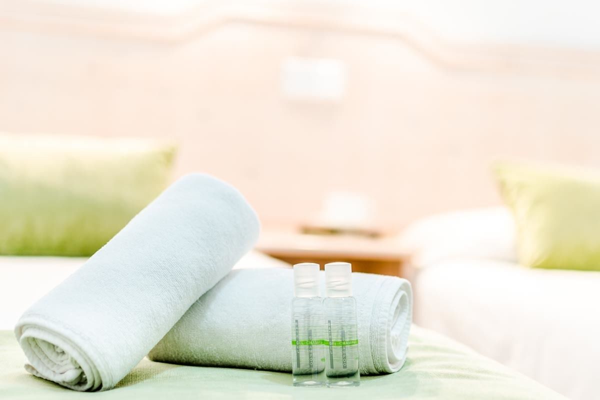 Una cama bien hecha con una toalla blanca enrollada y dos botellas transparentes con etiquetas verdes sobre una superficie verde.